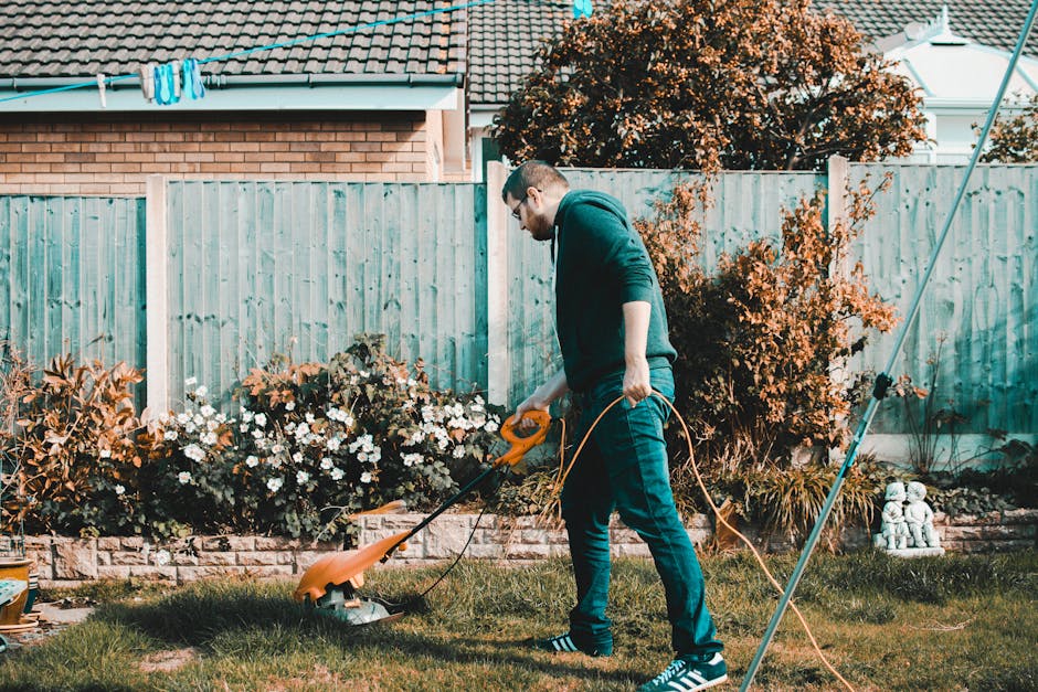 A young man wearing glasses, a dark hoodie, and jeans is lawn mowing a neatly maintained garden with a push lawn mower. The garden features a lush green lawn, bordered by flowerbeds filled with bloomi
